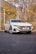 A sleek, white Volkswagen car with an elegant design is parked on a smooth asphalt surface. The vehicle has modern alloy wheels and a custom license plate reading 'STABLE'. Surrounding the car are autumn trees with yellow and brown leaves, and a large building backdrop.