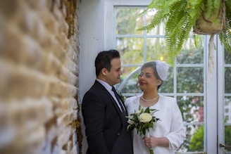 A couple is standing indoors with a soft focus on a brick wall to the left. The woman, holding a bridal bouquet, is wearing a white outfit with a small netted fascinator on her head and is looking at the man. The man is dressed in a dark suit and is gazing at her. There is a large window behind them showing a blurred outdoor scene with trees, and a hanging plant adds greenery to the setting.