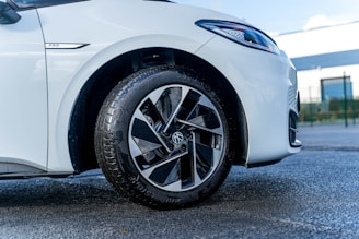 A close-up view of the front left wheel of a white car. The focus is on the tire and alloy wheel with distinct black and silver geometric patterns. The car is parked on a textured asphalt surface, and the background includes a blurred view of a building and a fence.
