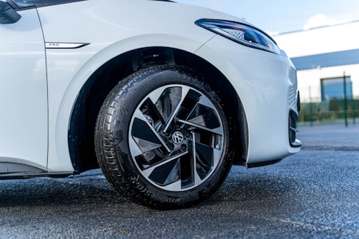 A close-up view of the front left wheel of a white car. The focus is on the tire and alloy wheel with distinct black and silver geometric patterns. The car is parked on a textured asphalt surface, and the background includes a blurred view of a building and a fence.
