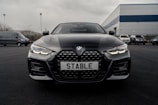 A sleek black BMW car is parked on a dark asphalt driveway with its front grille and headlights prominently displayed. The license plate reads 'STABLE'. The background features a few more vehicles and an industrial building with a blue and gray exterior.