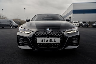 A sleek black BMW car is parked on a dark asphalt driveway with its front grille and headlights prominently displayed. The license plate reads 'STABLE'. The background features a few more vehicles and an industrial building with a blue and gray exterior.