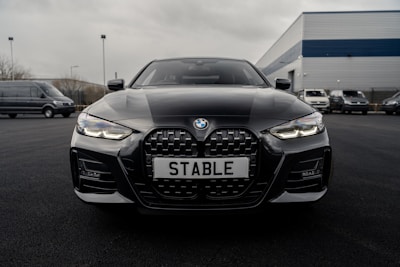 A sleek black BMW car is parked on a dark asphalt driveway with its front grille and headlights prominently displayed. The license plate reads 'STABLE'. The background features a few more vehicles and an industrial building with a blue and gray exterior.