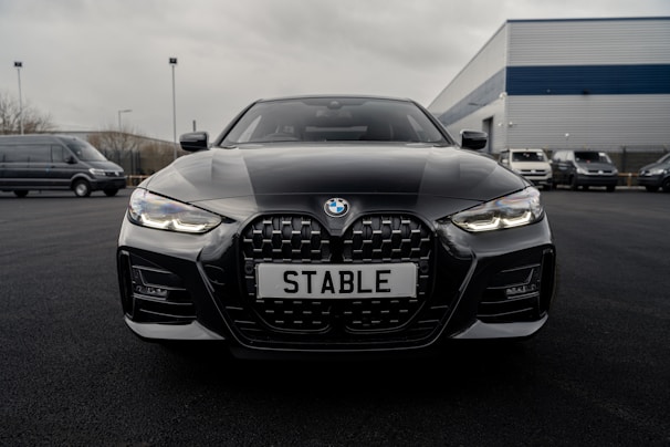 A sleek black BMW car is parked on a dark asphalt driveway with its front grille and headlights prominently displayed. The license plate reads 'STABLE'. The background features a few more vehicles and an industrial building with a blue and gray exterior.