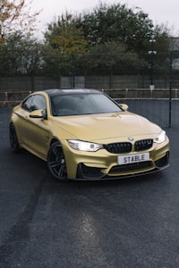 The image features a yellow BMW sports car parked on a wet asphalt surface. The car has a sleek and modern design with a prominent front grille adorned with the BMW logo. The license plate reads 'STABLE.' The setting appears to be an outdoor parking area with trees and a fence in the background.