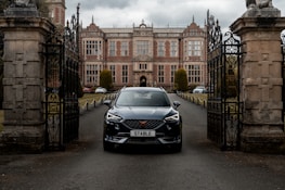 A sleek black car with the license plate 'STABLE' is prominently positioned in front of the entrance gates of an elegant, historical mansion. The gates are intricately designed with wrought iron, and the driveway leading to the mansion is lined with hedges and bollards. The mansion features a symmetrical facade with tall windows and a mix of stone and brick architecture.