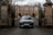 A sleek black car with the license plate 'STABLE' is prominently positioned in front of the entrance gates of an elegant, historical mansion. The gates are intricately designed with wrought iron, and the driveway leading to the mansion is lined with hedges and bollards. The mansion features a symmetrical facade with tall windows and a mix of stone and brick architecture.