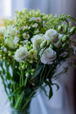 A bouquet of white roses and small daisies surrounded by green foliage, set against a softly blurred background. The flowers are in full bloom, adding a sense of freshness and elegance.