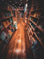 A cozy bookstore with wooden shelves filled with colorful books on both sides of a narrow aisle. The warm lighting casts a welcoming glow on the polished wooden floor, creating a comfortable and inviting atmosphere. The shelves hold a variety of books with different covers and sizes, suggesting a diverse selection available for browsing.