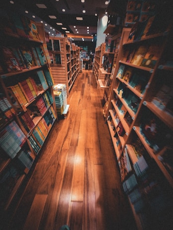 A cozy bookstore with wooden shelves filled with colorful books on both sides of a narrow aisle. The warm lighting casts a welcoming glow on the polished wooden floor, creating a comfortable and inviting atmosphere. The shelves hold a variety of books with different covers and sizes, suggesting a diverse selection available for browsing.