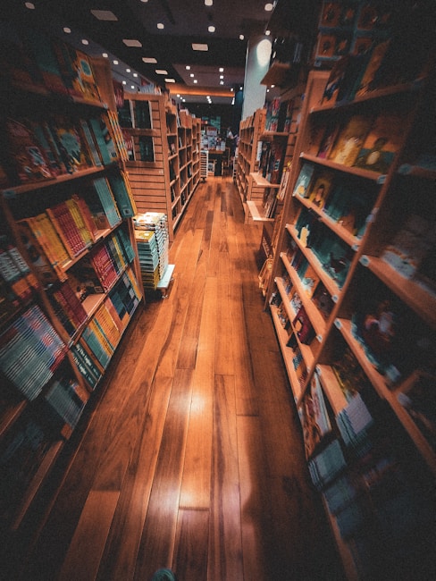 A cozy bookstore with wooden shelves filled with colorful books on both sides of a narrow aisle. The warm lighting casts a welcoming glow on the polished wooden floor, creating a comfortable and inviting atmosphere. The shelves hold a variety of books with different covers and sizes, suggesting a diverse selection available for browsing.