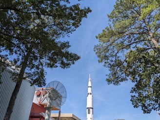 A tall white rocket stands prominently against a clear blue sky, surrounded by lush green trees. In the foreground, part of a large satellite dish is visible near industrial buildings with a corrugated metal siding.
