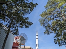 A tall white rocket stands prominently against a clear blue sky, surrounded by lush green trees. In the foreground, part of a large satellite dish is visible near industrial buildings with a corrugated metal siding.