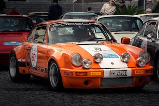 A vintage orange and white sports car is parked among other classic cars at a vehicle gathering or rally. The car is adorned with racing decals and the number 80 displayed prominently on its door. Several auxiliary headlights are mounted on the front bumper, adding to its race-ready appearance. The surrounding environment features cobblestone paving and other classic cars, with people standing nearby, suggesting a show or exhibition setting.