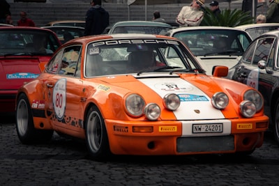 A vintage orange and white sports car is parked among other classic cars at a vehicle gathering or rally. The car is adorned with racing decals and the number 80 displayed prominently on its door. Several auxiliary headlights are mounted on the front bumper, adding to its race-ready appearance. The surrounding environment features cobblestone paving and other classic cars, with people standing nearby, suggesting a show or exhibition setting.