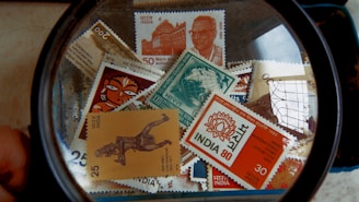 A collection of Indian postal stamps viewed through a magnifying glass. The stamps feature various designs, including architectural landmarks, artwork, cultural symbols, and portraits. The magnifying glass emphasizes the details and intricacies of the stamp designs.