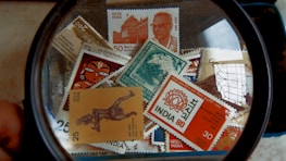 A collection of Indian postal stamps viewed through a magnifying glass. The stamps feature various designs, including architectural landmarks, artwork, cultural symbols, and portraits. The magnifying glass emphasizes the details and intricacies of the stamp designs.