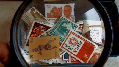 A collection of Indian postal stamps viewed through a magnifying glass. The stamps feature various designs, including architectural landmarks, artwork, cultural symbols, and portraits. The magnifying glass emphasizes the details and intricacies of the stamp designs.