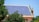 A rooftop is covered with an array of blue solar panels, surrounded by lush green trees. The building is constructed with red bricks and has a classic architectural style. The sky is clear, enhancing the overall brightness.