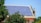 A rooftop is covered with an array of blue solar panels, surrounded by lush green trees. The building is constructed with red bricks and has a classic architectural style. The sky is clear, enhancing the overall brightness.
