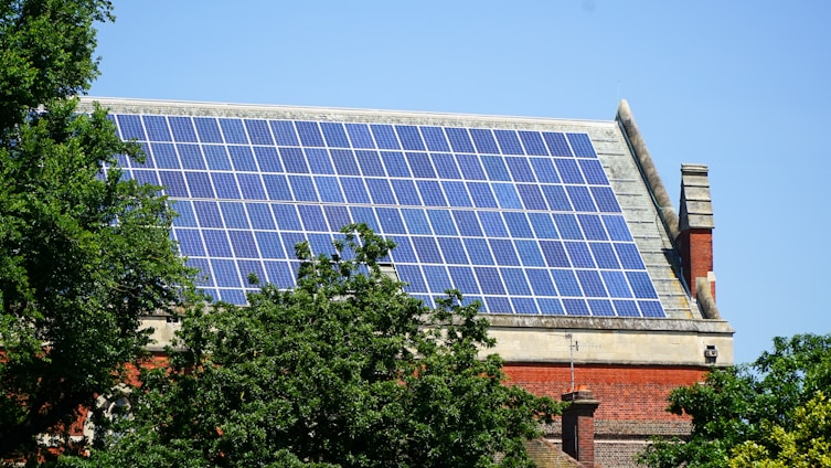 A rooftop is covered with an array of blue solar panels, surrounded by lush green trees. The building is constructed with red bricks and has a classic architectural style. The sky is clear, enhancing the overall brightness.
