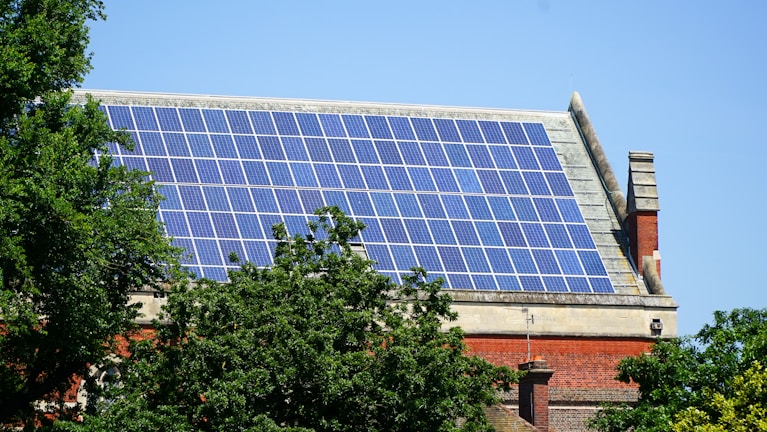 A rooftop is covered with an array of blue solar panels, surrounded by lush green trees. The building is constructed with red bricks and has a classic architectural style. The sky is clear, enhancing the overall brightness.