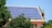 A rooftop is covered with an array of blue solar panels, surrounded by lush green trees. The building is constructed with red bricks and has a classic architectural style. The sky is clear, enhancing the overall brightness.