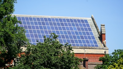 A rooftop is covered with an array of blue solar panels, surrounded by lush green trees. The building is constructed with red bricks and has a classic architectural style. The sky is clear, enhancing the overall brightness.