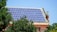 A rooftop is covered with an array of blue solar panels, surrounded by lush green trees. The building is constructed with red bricks and has a classic architectural style. The sky is clear, enhancing the overall brightness.