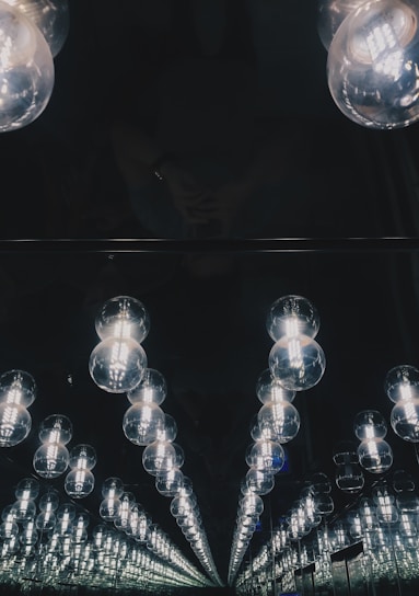 An array of illuminated glass bulbs is reflected infinitely in a mirror, creating a tunnel-like visual effect. The lighting is dim, with a focus on the bright bulbs against a dark background.