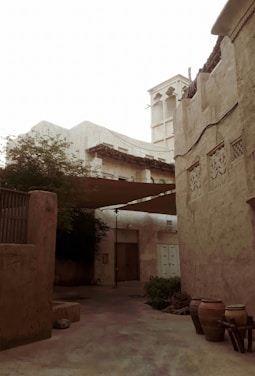 An old, rustic courtyard with high walls and intricate designs on the plaster. There is a traditional wind tower structure, commonly used for natural ventilation. The space is shaded with cloth and has various clay pots, some greenery, and a wooden door. The overall architecture suggests an historical or cultural heritage setting.