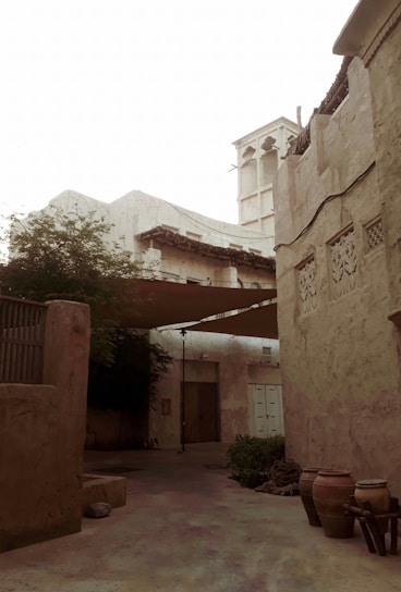 An old, rustic courtyard with high walls and intricate designs on the plaster. There is a traditional wind tower structure, commonly used for natural ventilation. The space is shaded with cloth and has various clay pots, some greenery, and a wooden door. The overall architecture suggests an historical or cultural heritage setting.