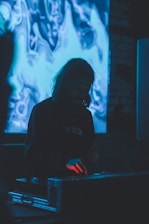 A person with long hair stands in a dimly lit environment, interacting with equipment that emits a soft blue glow. The background features an abstract display that appears to be a digital screen with swirling blue and white patterns.