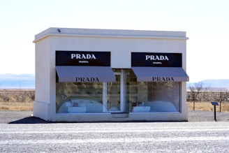 A small white building resembling a retail storefront with large glass windows and two black awnings displaying the Prada logo. The interior appears unfinished, with shelves holding a few handbags. Situated in an isolated, arid landscape with a clear blue sky in the background.