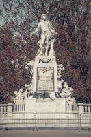A grand white statue of a historical figure stands atop an intricately decorated base, surrounded by cherubic figures. The name 'Mozart' is prominently displayed on the central plaque, surrounded by musical motifs and laurel garlands. The background features dense trees with autumnal red foliage.