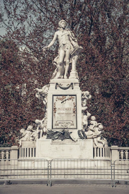 A grand white statue of a historical figure stands atop an intricately decorated base, surrounded by cherubic figures. The name 'Mozart' is prominently displayed on the central plaque, surrounded by musical motifs and laurel garlands. The background features dense trees with autumnal red foliage.
