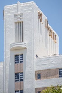 A white Art Deco building with architectural features such as geometric lines and decorative elements. The structure has several windows with metal frames, and the facade includes vertical and horizontal grooves, giving it a streamlined appearance. The light plays on its textured surface under a clear blue sky.