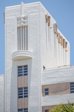 A white Art Deco building with architectural features such as geometric lines and decorative elements. The structure has several windows with metal frames, and the facade includes vertical and horizontal grooves, giving it a streamlined appearance. The light plays on its textured surface under a clear blue sky.