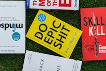 Several books placed on a grass surface, including titles like 'Mindsight,' 'Do Epic Shit,' and 'Skill Kill.' The covers of the books are visible, with varying colors including yellow, red, and blue. The books are arranged in a casual, overlapping manner.