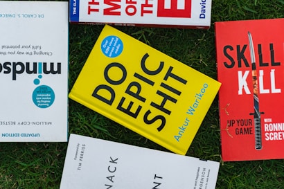 Several books placed on a grass surface, including titles like 'Mindsight,' 'Do Epic Shit,' and 'Skill Kill.' The covers of the books are visible, with varying colors including yellow, red, and blue. The books are arranged in a casual, overlapping manner.