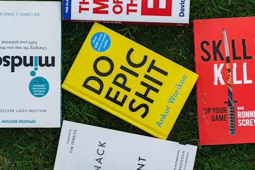 Several books placed on a grass surface, including titles like 'Mindsight,' 'Do Epic Shit,' and 'Skill Kill.' The covers of the books are visible, with varying colors including yellow, red, and blue. The books are arranged in a casual, overlapping manner.
