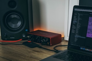 A computer setup featuring a laptop with audio editing software open, connected to an audio interface. There is a speaker on the left side, and cables are visibly plugged into the devices. The background is a wall with subtle lighting creating a warm ambiance.