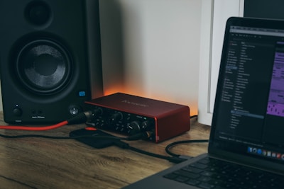 A computer setup featuring a laptop with audio editing software open, connected to an audio interface. There is a speaker on the left side, and cables are visibly plugged into the devices. The background is a wall with subtle lighting creating a warm ambiance.