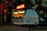 A vintage-style, dome-shaped food cart with a futuristic design is situated on a paved path. The cart is lit up with bright neon lights, featuring signage in a foreign script. It has a retro, space-themed appearance with a small service window displaying various items. Surrounding the cart are trees and buildings, with dim ambient lighting creating a cozy evening atmosphere.