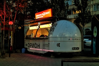 A vintage-style, dome-shaped food cart with a futuristic design is situated on a paved path. The cart is lit up with bright neon lights, featuring signage in a foreign script. It has a retro, space-themed appearance with a small service window displaying various items. Surrounding the cart are trees and buildings, with dim ambient lighting creating a cozy evening atmosphere.