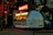 A vintage-style, dome-shaped food cart with a futuristic design is situated on a paved path. The cart is lit up with bright neon lights, featuring signage in a foreign script. It has a retro, space-themed appearance with a small service window displaying various items. Surrounding the cart are trees and buildings, with dim ambient lighting creating a cozy evening atmosphere.