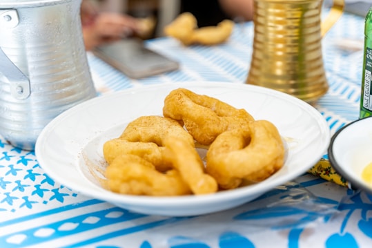 A white plate holds several pieces of fried dough, which are golden brown in color. The table is covered with a blue and white patterned cloth. Surrounding the plate are metal containers, a green bottle, and a smartphone. The setting appears casual and possibly outdoors.