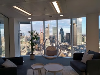 A modern office space with large floor-to-ceiling windows offering a panoramic view of a city skyline, including several skyscrapers. The interior features contemporary furniture including dark sofas, a round glass hanging chair, and a small table. A potted plant adds a natural element near the window.