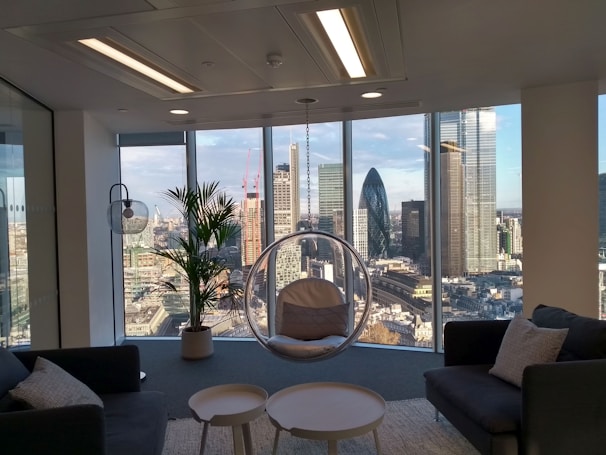 A modern office space with large floor-to-ceiling windows offering a panoramic view of a city skyline, including several skyscrapers. The interior features contemporary furniture including dark sofas, a round glass hanging chair, and a small table. A potted plant adds a natural element near the window.