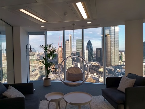 A modern office space with large floor-to-ceiling windows offering a panoramic view of a city skyline, including several skyscrapers. The interior features contemporary furniture including dark sofas, a round glass hanging chair, and a small table. A potted plant adds a natural element near the window.